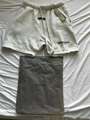 Essentials Fear of God shorts grå - Gråa shorts från Essentials Fear of God med svart logga på vänster ben. Mjuk bomullsblandning, elastisk midja med snörning och avslappnad passform. Perfekta för sommaren och chill dagar.