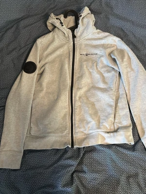 Grå hoodie från Sail Racing - Snygg grå hoodie från Sail Racing med dragkedja, huva och svart logga på bröstet. Stor text 'SAIL RACING ANTARCTICA EXPEDITION' på ryggen och en svart patch på ena ärmen. Tillverkad i mjukt material, perfekt för chill dagar eller när du vill ha en sportig look.