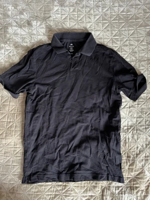 Svart pikétröja från H&M - Klassisk svart pikétröja från H&M med regular fit. Tröjan har korta ärmar, krage och knappar framtill. 