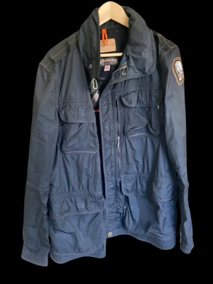 Mörkblå vindjacka från Parajumpers - Snygg mörkblå vindjacka från Parajumpers med flera praktiska fickor framtill, hög krage och patch på ärmen. Jackan har dragkedja, tryckknappar och detaljer i orange. Tillverkad i lätt och slitstarkt material, perfekt för blåsiga dagar. Har används väldigt lite och framstår därför som ny, Liten i storleken och passar snarare någon som är L-XL än XXL.. 