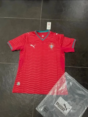 Portugal röd matchtröja från Puma - Snygg röd Portugal matchtröja från Puma med vita vågmönster, gröna detaljer vid krage och ärmslut samt Portugal-emblem på bröstet. Tröjan har korta ärmar och sportig passform, perfekt för fotboll eller träning.