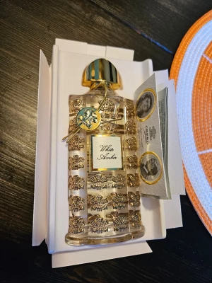 Creed White Amber parfym 75 ml - Lyxig parfym från Creed, White Amber, i en exklusiv och elegant genomskinlig flaska med gulddetaljer och dekorativa mönster. Flaskan har en guldfärgad kork och levereras i en snygg vit förpackning med gulddetaljer. Innehåll: 75 ml. Obs! Ej äkta.