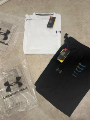 Under Armour träningsset t-shirt & shorts - Snyggt träningsset från Under Armour med en vit kortärmad t-shirt och svarta shorts. T-shirten har logga på bröstet och sidan, och shortsen har dragsko i midjan samt logga och tryck i färg. Perfekt för gymmet eller löpning.