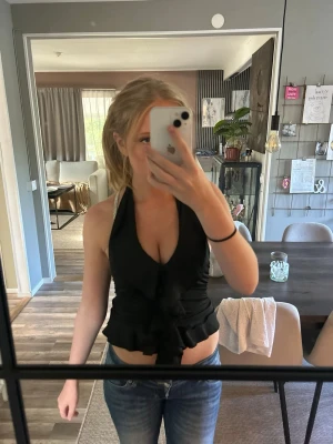 Svart omlott halterneck topp - Snygg svart omlott halterneck topp med knyt i ryggen och volangkant nedtill. Toppen är croppad och har en djup v-ringning framtill. Perfekt för dig som vill ha en trendig och stilren look.