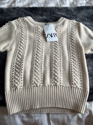 Beige stickad topp från Zara - Snygg beige stickad topp från Zara med korta ärmar och coolt strukturmönster i lodräta ränder. Tröjan har ribbstickad nederkant och halsringning. Perfekt för dig som gillar en stilren och trendig look.