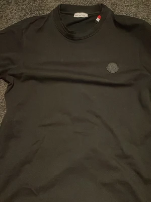 Svart Moncler t-shirt med logga - Snygg svart t-shirt från Moncler med diskret svart logga på bröstet och klassisk rund hals. T-shirten har korta ärmar och är gjord i mjuk bomull som känns skön mot huden. Perfekt för dig som gillar stilrena och exklusiva plagg. Dubbel krage för en speciel stil, perfekt till sommar outfits och utekvällar. FINNS I VIT OCKSÅ!