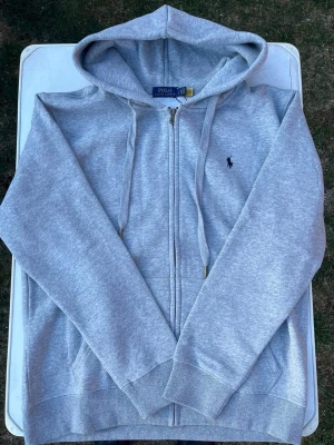 Grå zip hoodie från Polo Ralph Lauren - Snygg grå hoodie med dragkedja från Polo Ralph Lauren. Klassisk design med huva, dragsnören och fickor framtill. Ikoniska mörkblå polospelaren broderad på bröstet. Tillverkad i mjuk bomullsblandning för skön känsla.