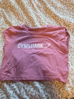 Rosa croppad Gymshark t-shirt - Snygg rosa croppad t-shirt från Gymshark med vitt tryck framtill. T-shirten har korta ärmar och är tillverkad i mjuk bomull, perfekt för träning eller chill. Enkel och sportig design med rund hals.