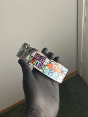 Vit Bälte med mönster  -  skärp med graffiti-inspirerat tryck i flera färger och stor -logga i silver. Bältet har en vit bas med detaljer i blått, orange, grönt och lila. Snygg accessoar för dig som vill sticka ut.