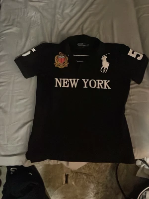 Svart pikétröja från Ralph Lauren - Svart pikétröja från Ralph Lauren med vit logga, texten 'NEW YORK' på bröstet och siffran 5 på ärmen och ryggen. Tröjan har klassisk krage och korta ärmar, samt broderad detalj på bröstet.