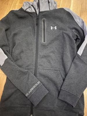 Mörkgrå hoodie från Under Armour - Snygg mörkgrå hoodie från Under Armour med dragkedja, huva och kontrasterande ljusgrå detaljer på axlar och huva. Bröstficka med dragkedja och logga på bröstet samt ärmen. 