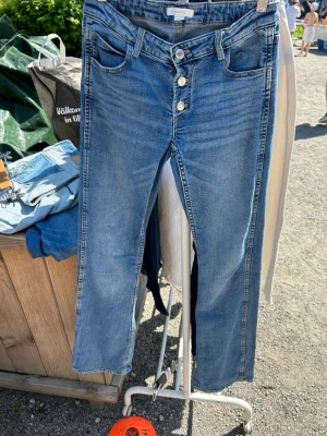 Blå bootcut jeans från Gina Tricot - Snygga blå jeans från Gina Tricot med utsvängda och låg midja. Jeansen har fyra synliga knappar framtill, klassiska fem fickor och bälteshällor. Tillverkade i mjuk denim med lätt tvättad look som ger en avslappnad vibe.