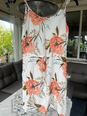 Vit blommig sommarklänning med knyt - Säljer en vit klänning med stora rosa blommor och gröna blad. Klänningen har smala axelband, knyt i midjan och är i en luftig modell som passar perfekt till sommaren. Materialet känns lätt och svalt. Den har dock en fläck på framsidan, men som knappt syns
