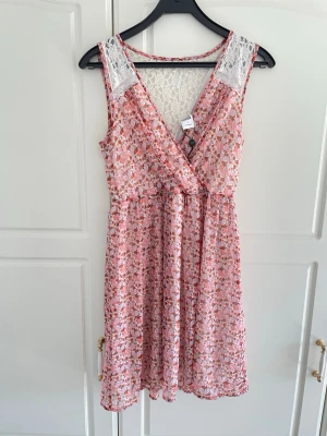 Blommig omlottklänning Vero Moda - Söt blommig kortklänning från Vero Moda i rosa och vita toner. Klänningen har omlottmodell, spetsdetaljer vid axlar och rygg samt v-ringning. Lätt och luftig med ett mjukt fall, perfekt för varma dagar.