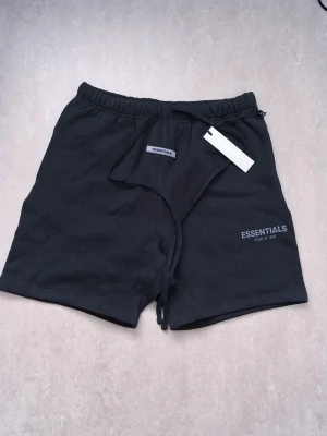 Svarta shorts från Essentials Fear of God - Svarta shorts från Essentials Fear of God med elastisk midja och snörning. Klassisk passform med en bakficka och logga framtill. Perfekta för en avslappnad stil och tillverkade i mjuk bomull.