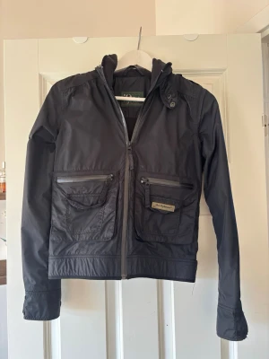 Svart vindjacka Peak Performance - Snygg svart vindjacka från Peak Performance med två stora bröstfickor med dragkedja och lock. Jackan har hög krage med knappdetalj och är tillverkad i ett lätt, vindtätt material. Perfekt för blåsiga dagar och enkel att matcha med olika outfits.