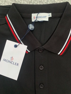 Svart pikétröja från Moncler - Snygg svart pikétröja från Moncler med klassisk krage och knappar framtill. Kragen har röda och vita ränder som ger en sportig touch. Tröjan har Moncler-logga broderad på bröstet och är tillverkad i bomull.