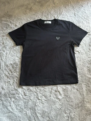 Svart t-shirt Comme des Garçons Play - Svart t-shirt från Comme des Garçons Play med det ikoniska hjärte-emblemet broderat på bröstet. Klassisk rund halsringning och korta ärmar. Tillverkad i mjuk bomull som känns skön mot huden. Perfekt för en clean och stilren look.