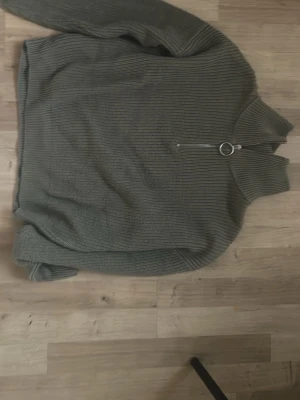 Grå ribbstickad tröja med dragkedja - Mysig grön stickad tröja med hög krage och halv dragkedja med ringdetalj. Tröjan har ribbstickat mönster och långa ärmar, perfekt för kyliga dagar. Passar dig som gillar enkel och stilren design.