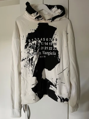Maison Margiela vit hoodie med tryck - Säljer en vit hoodie från Maison Margiela med svart abstrakt tryck och logga på bröstet. Hoodien har huva och ribbade muddar. Oversized fit och cool streetwear-vibe. Materialet känns som mjuk bomull och trycket ger en unik look.