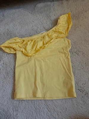 Gul oneshoulder topp från Kappahl - Snygg gul oneshoulder topp från Kappahl med volangdetalj längs halsringningen. Tillverkad i mjuk bomull, perfekt för varma dagar. Enkel men trendig design som sticker ut med sin asymmetriska axel.