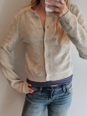Beige kofta från H&M Divided - Mysig beige kofta från H&M Divided med rund halsringning och knappar framtill. Koftan är i mjukt material och har långa ärmar samt ribbade muddar vid ärmslut och nederkant. Perfekt att slänga över en topp om det är lite kyligt ute.