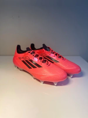 Adidas F50 Elite (SG) - Säljer ett nytt par Adidas F50 elite fotbollsskor i en snygg rosa färg med svarta detaljer och klassiska tre ränder på sidan. Skorna är aldrig använda och har dobbar i metall för bästa grepp på planen och är tillverkade i ett lätt syntetmaterial för snabbhet och smidighet. 