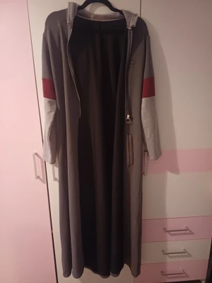  hooded loungewear  - Mysig lång loungewear i grått med huva och dragkedja framtill. Ärmarna har ljusgrå partier och en bred röd rand över armbågarna. Perfekt för chill dagar hemma. Materialet känns mjukt och bekvämt, troligen bomullsblandning.