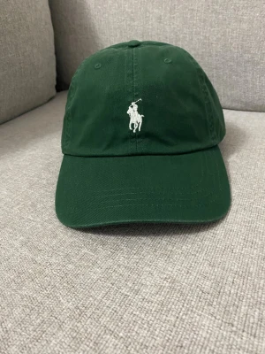 Mörkgrön keps från Polo Ralph Lauren - Snygg mörkgrön keps från Polo Ralph Lauren med klassisk böjd skärm och justerbart spänne bak. Framtill finns den ikoniska broderade polospelaren i vitt och baktill står det 'POLO' broderat. Tillverkad i bomull för en skön och avslappnad look.