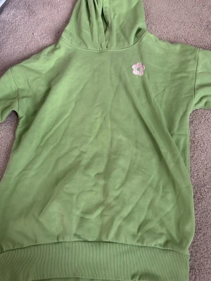 Grön hoodie med lång ärm och blomma - Säljer en ljusgrön hoodie med långa ärmar och huva. På bröstet finns en liten broderad blomma i rosa och vitt. Hoodien har ribbade muddar nedtill och är gjord i mjukt bomullsliknande material. Perfekt för en chill och avslappnad stil.