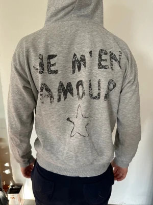 Grå hoodie med tryck och huva - Snygg grå hoodie med huva och tryck i svart text på ryggen: 'JE M'EN AMOUR' samt en stjärna. På bröstet står det 'MA CHERIE' och en mindre stjärna. Hoodien har dragsko i huvan och är gjord i mjukt sweatshirtmaterial.