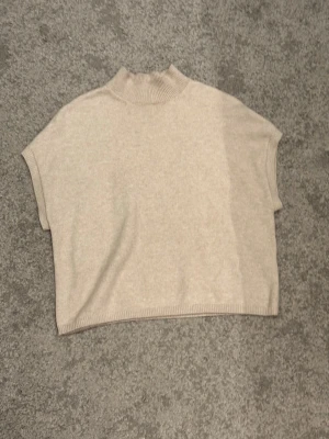 Beige stickad kortärmad topp - Snygg beige stickad topp med korta ärmar och hög krage. Perfekt att styla över en långärmad tröja eller bära som den är. Mjuk och skön kvalitet med ribbad kant vid hals och nederkant. Enkel och clean design som passar till mycket.