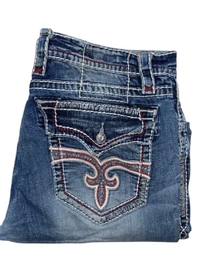 Blåa jeans från Rock Revival - Snygga blå jeans från Rock Revival med unika broderade bakfickor och kontrastsömmar. Jeansen har en klassisk femficksmodell, raka ben och slitna detaljer framtill. Materialet är denim i bomull med cool tvätt och dekorativa knappar. (Ena fickknappen är borta och de finns lite lagningar men de är inget man märker och vissa tycker det är snyggt)