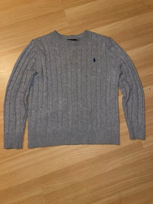 Grå stickad polo Ralp Lauren - Kabelstickad grå tröja från Polo Ralph Lauren med rund halsringning och den klassiska mörkblå logotypen broderad på bröstet. Tröjan har långa ärmar och ribbade muddar vid ärmslut och nederkant. Perfekt för en stilren och avslappnad look. PRIS KAN DISKUTERAS 