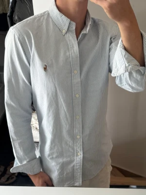 Randig skjorta från Ralph Lauren - Klassisk skjorta från Ralph Lauren med tunna blå och vita ränder. Skjortan har button-down krage, långa ärmar och knappar framtill. Broderad logga på bröstet ger en stilren touch. Perfekt till jeans eller chinos för en clean look.