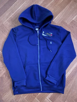 Marinblå hoodie från Polo Ralph Lauren - Snygg marinblå hoodie från Polo Ralph Lauren med dragkedja framtill och klassisk vit logga broderad på bröstet. Hoodien har huva med dragsko och ribbade muddar vid ärmar och nederkant. Tillverkad i mjukt bomullsmaterial för skön känsla.