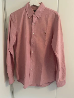 Rosa skjorta från Polo Ralph Lauren - Klassisk rosa skjorta från Polo Ralph Lauren med button-down krage och broderad logga på bröstet. Skjortan har långa ärmar och vita knappar framtill. Perfekt för en stilren och avslappnad look. Pyttelitet hål som är lagat på ena ärmen, det syns inte alls när man har på sig skjortan eftersom det ligger på undersidan av armen, se sista bild.