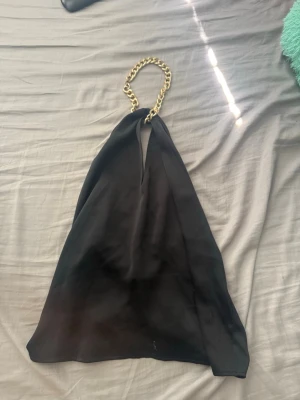 Svart halterneck topp med guldkedja - Snygg svart halterneck topp med bred guldfärgad kedja som halsband. Toppen har en djup v-ringning och är ärmlös med en lös passform. Använd 1 gång 💕