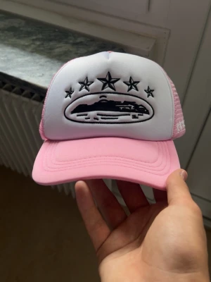 Rosa och vit truckerkeps med brodyr - Säljer en snygg truckerkeps i rosa och vitt med mesh på sidorna och broderat UFO-motiv med stjärnor framtill. Justerbar baktill för perfekt passform. Perfekt accessoar för dig som vill sticka ut med en unik och trendig keps.