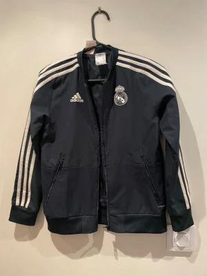 Mörkblå Real Madrid adidas jacka - Snygg mörkblå träningsjacka från adidas med Real Madrid-logga på bröstet och klassiska vita ränder längs ärmarna. Jackan har dragkedja framtill, två fickor med dragkedja och ribbade muddar. Perfekt för sport eller chill.