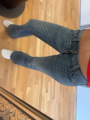 Blå bootcut jeans med låg midja - Säljer ett par blå bootcut jeans med låg midja och klassisk femficksmodell. Jeansen har snygga slitningar och en lätt tvättad look. Perfekta för dig som gillar retrostil och vill ha ett par jeans som sticker ut lite extra.