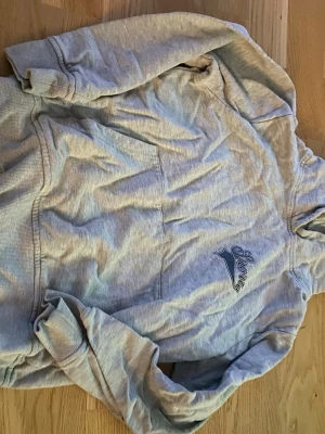 Grå hoodie med Shorty-tryck - Snygg grå hoodie med huva och känguruficka framtill. På bröstet finns ett tryck med texten 'Shorty' i mörkblått. Hoodien är gjord i mjukt bomullsmaterial och har ribbade muddar vid ärmar och nederkant. Perfekt för en avslappnad och chill stil.