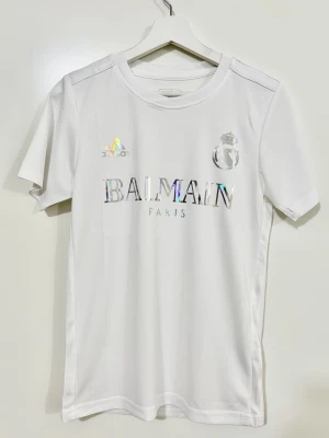 Vit Real Madrid Balmain x Adidas t-shirt  - Snygg vit t-shirt med holografiskt Balmain Paris-tryck på bröstet och Adidas-logga. T-shirten har korta ärmar och rund hals. Materialet känns som polyester, vilket gör den lätt och bekväm. Perfekt för dig som gillar stilrena och exklusiva plagg.