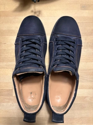 Blå Loubs - Snygga mörkblå sneakers från Louboutin i blå mocka med matchande mörkblå snörning och en röd klassisk sula. Skorna har en låg profil och stilren design som passar till det mesta. Insidan är ljusbeige och de har en enkel pull-tab bak för lättare påtagning.