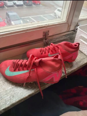 Röda Nike Mercurial fotbollsskor - Säljer ett par röda Nike Mercurial fotbollsskor med turkos swoosh och detaljer. Skorna har snörning och en strumpliknande krage för extra stöd runt ankeln. Tillverkade i syntetmaterial för bra grepp och kontroll på planen. Perfekta för dig som vill sticka ut på matchen.