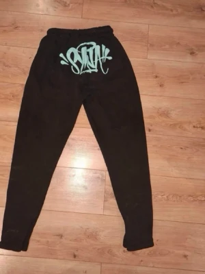 Svarta mjukisbyxor med turkost syna world tryck - Säljer ett par svarta mjukisbyxor med syna world märket i turkos. Byxorna har resår i midjan och vid bensluten för skön passform. Perfekta för chill dagar eller streetwear-stil. Materialet känns mjukt och bekvämt är i storlek L och original paketet kommer med dock så råkade jag öppna orginal pake  på fel sätt men annars är allt som orginalt och inget är sönder och jag kan skicka mer bilder och svara på frågor 