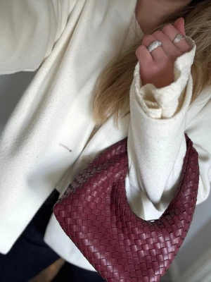 Vinröd flätad handväska  - Säljer en vinröd handväska med flätad design och mjuk, böjd form, tillverkad i skinnliknande material. Tipsar om att lägga i ett matlåde-lock eller en kartong byt i bottnen för att få den platta looken om man föredrar den istället för den lite böjda looken.