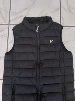 Svart dunväst från Lyle & Scott - Snygg svart dunväst från Lyle & Scott med gul log- broderad på bröstet. Västen har hög krage, just sop. bricks och två dragkedjeförsedda fickor. Perfekt för lager-på-lager och håller också värmen med sin vadderade design. De ända täknet på användning är en liten svart klister bit som sitter på framsidan av västen. Den sitter där för den fick en mindre skada. Ni kan se det på den sista bilden. 