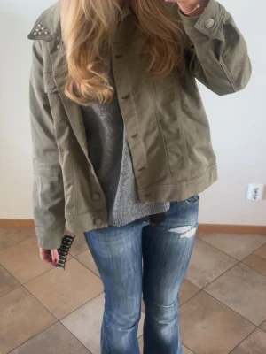 Olivgrön jeansjacka med nitar - Snygg olivgrön jeansjacka med coola nitar på axlarna. Klassisk modell med knappar framtill och raka sömmar. Perfekt att slänga över en hoodie eller tröja för en avslappnad look.
