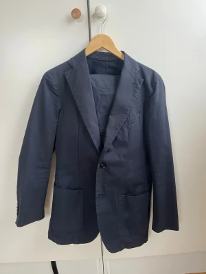 Casual kostym från SUITSUPPLY  - Stilren mörkblå kavaj med två stora patchfickor framtill och klassiska slag. Kavajen har dubbelknäppning , perfekt för en avslappnad men ändå uppklädd look. Passar till de fina tillställningarna men går utmärkt till en vit T-shirt och sneakers nu i vår och sommar. Bra snygg passform. Nypris 5900 kr köpt i stockholm